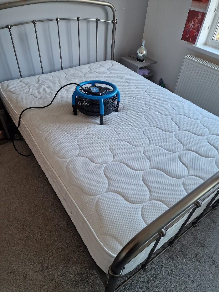 Blue portable air blower sits on a white quilted mattress in a bedroom, plugged in by the head of the bed.