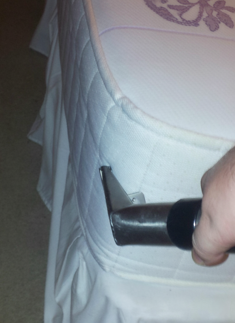 Close-up of a hand gripping a metal tool with a flat angled blade being inserted into the edge of a white quilted mattress.