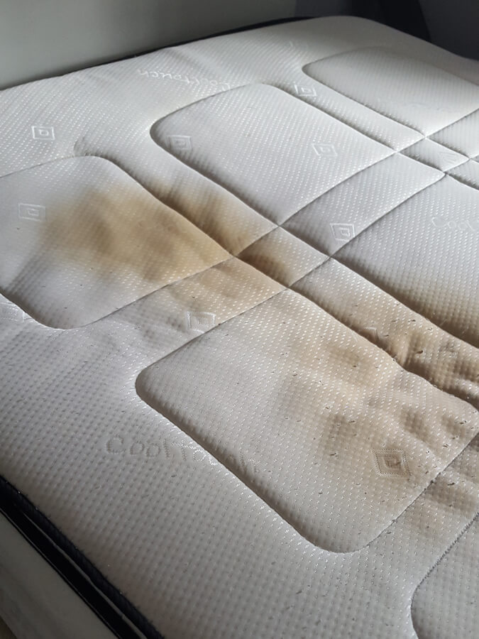Close-up of a light-colored quilted mattress top with brown stains and patches of wear.